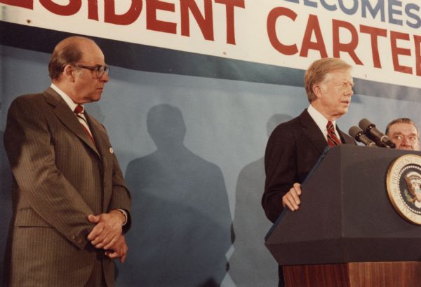 President Jimmy Carter standing behind a podium withGaylord Nelson on the left. Another man is behind the podium on the right. A banner hangs in the background.