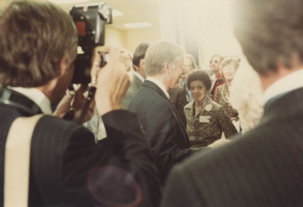 Vel Phillips is in the background looking at President Jimmy Carter standing among a crowd. There is a photographer in the foreground holding up a camera.