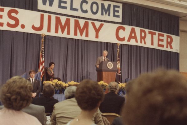 View from audience of President Jimmy Carter addressing a crowd of people from a stage. On the left Vel Phillips sits on the stage near an American Flag. A banner hangs in the background that reads: "Welcome Pres. Jimmy Carter."