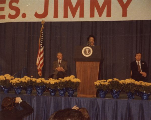 View from audience of Vel Phillips speaking on stage at a podium. On the left is President Jimmy Carter, who is wearing a suit and smiling at Vel. There are yellow flowers lining the front of the stage. Photographers are in the foreground.
