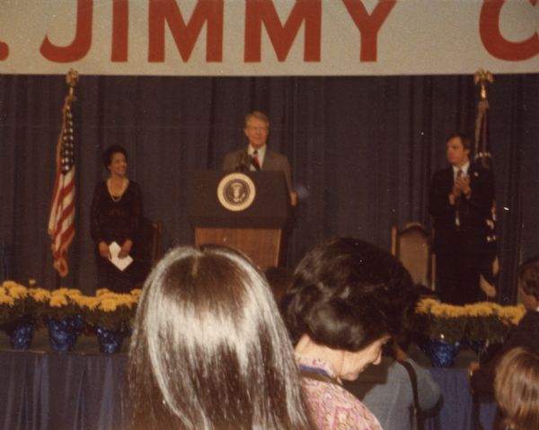 View from audience of President Jimmy Carter standing at a podium. Vel Phillips stands on the left. On the right is a man standing and applauding.