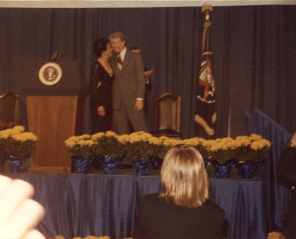 View from audience of Vel Phillips embracing President Jimmy Carter on a stage near a podium. A presidential flag is on the right of the stage.