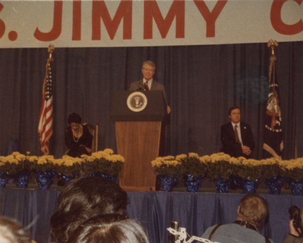 View from audience of President Jimmy Carter speaking at a podium. On the left Vel Phillips sits near an American Flag. A banner above reads: "Pres. Jimmy Carter."