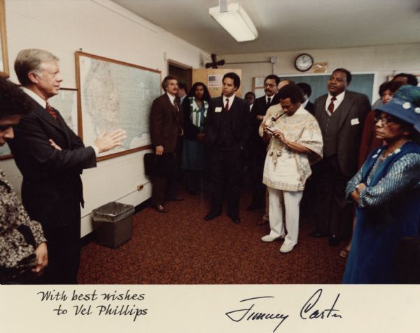 Former President Jimmy Carter addressing a crowd of people in a classroom. Vel Phillips is on the far left standing next to the President. A group of people stand listening in the room and in the open doorway.