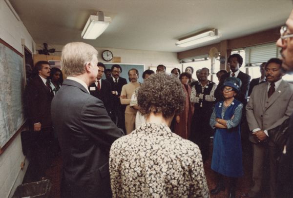 Rear view of President Jimmy Carter and Vel Phillips standing in a classroom talking with a group of people. There is a map on the left and a chalkboard in the background.