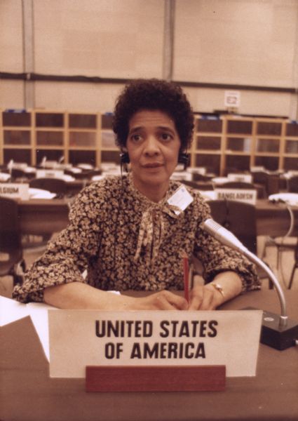 Vel Phillips in a floral top and wearing headphones and a name tag. She is sitting at a desk with a microphone and a sign that says "United States of America." Vel is attending the United Nations Decade for Women conference.