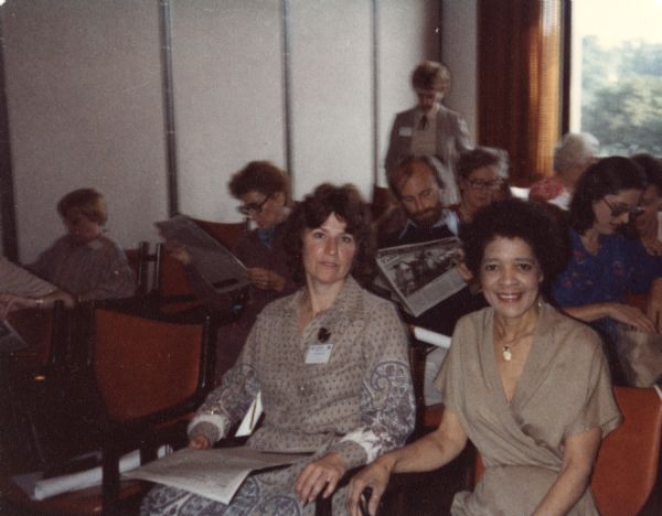 Vel Phillips wearing a light brown outfit and necklace. She is sitting next to another woman with brown hair. They are sitting in a waiting room with other people.