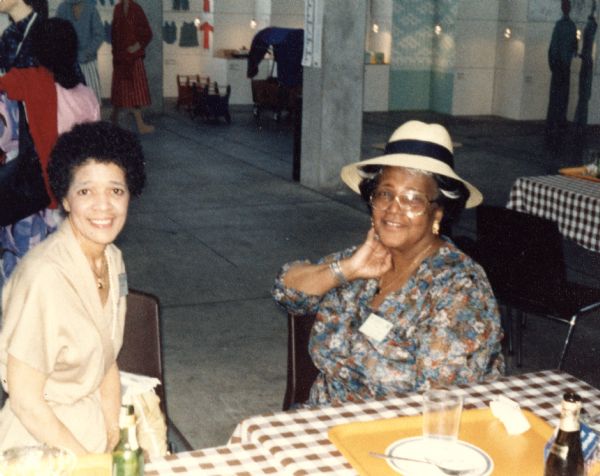 Vel Phillips wearing a tan outfit and sitting next to Anne Turpeau, who is wearing a floral top, hat and eyeglasses. There are sitting at tables with plaid tablecloths. Both women were United Nations delegates at the Decade for Women conference in Copenhagen.