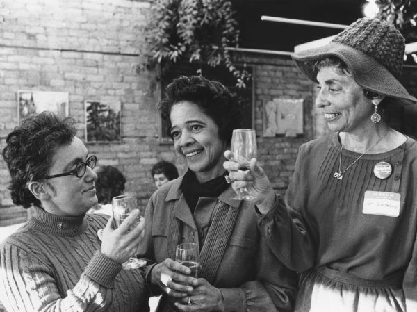 Vel Phillips stands between two women. They are all holding wine glasses. The woman on the left is wearing glasses and a turtleneck. The woman on the right is wearing a hat and an "ERA" necklace. In the background people sit at tables near a brick wall hung paintings.