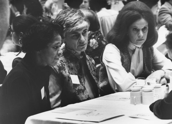 Vel Phillips, on the left, sitting with two other woman at a table. They are all wearing name tags. Other women sit at tables behind them.