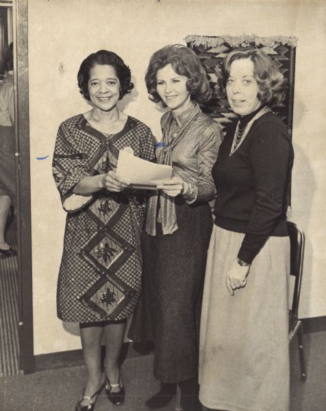 Vel Phillips stands in a room with two other women. Vel is wearing a buba and holding a document with the woman in the center. Vel had purchased the buba at an inner city arts fair and wore it to a council meeting in July 1969.