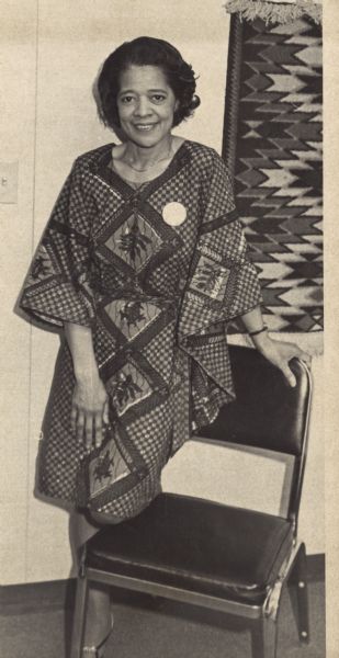 Vel Phillips in a buba, posing next to a chair. A textile print hangs on the wall behind her. Vel purchased the buba at an inner city arts fair and wore it to a council meeting in July 1969.
