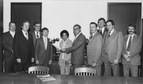 Group portrait of Vel Phillips in a room with nine men. She is standing in the center holding a document with a man wearing a suit. On the left are four men, and to the right are another four men. In the foreground is a desk with a magazine on it with the title "Wisconsin."