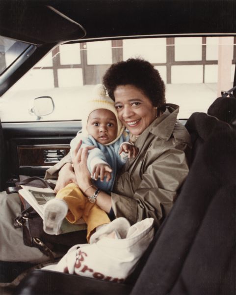 View across front seat towards Vel Phillips sitting in the passenger seat of a car holding a baby in her lap. Vel is wearing a coat and the baby is wearing a light blue coat and a yellow hat.