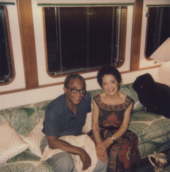 Vel Phillips and her husband W. Dale Phillips sitting on a green and white tree-patterned couch. Dale is wearing a blue shirt and eyeglasses. Vel is wearing a patterned dress.