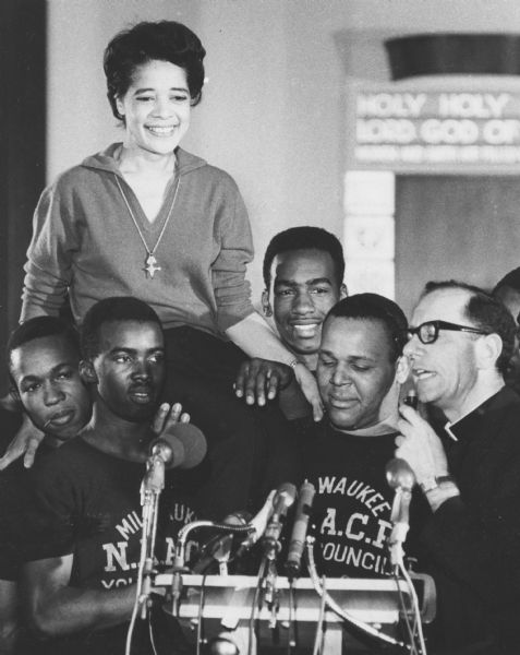 Vel Phillips being carried on the shoulders of Milwaukee NAACP Youth Council members. On the right is Father James Groppi speaking into a microphone on a podium with many microphones. In the background is a sign that reads, in part: "Holy Holy . . . Lord God of . . ."