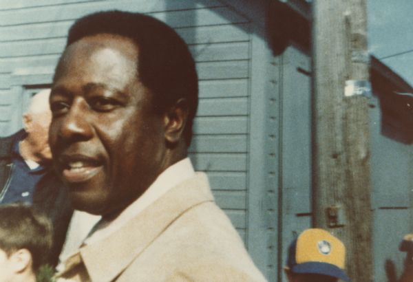 Head and shoulders portrait of Hank Aaron outdoors. Behind him are a man, and two children. The boy on the right is wearing a yellow and blue Milwaukee Brewers baseball hat.