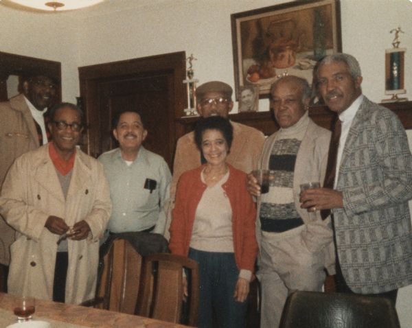 Vel Phillips standing for a group portrait indoors with six men. Second from the left is her husband W. Dale Phillips. There are trophies on a mantle in the background.