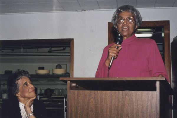 Vel Phillips sitting, on left, and looking on as Mother Naomi Scott speaks into a microphone at a podium at the Community Brainstorming Conference.