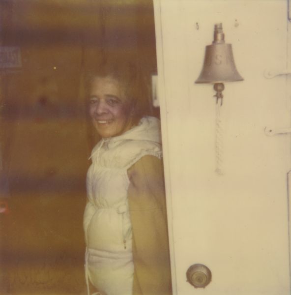 Vel Phillips, in a white vest, stand near an open doorway with a bell on it that is engranved with: "U.S."