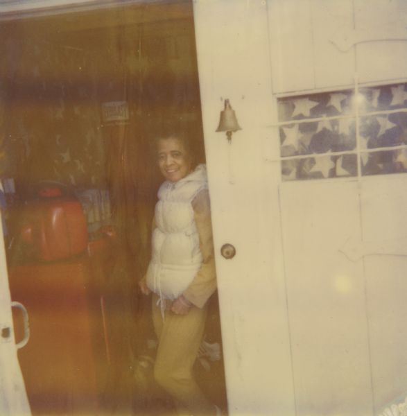 Vel Phillips standing in an open double doorway wearing a white puffy vest and yellow pants. A bell is hanging on the closed door, which has a window with star patterned curtains.