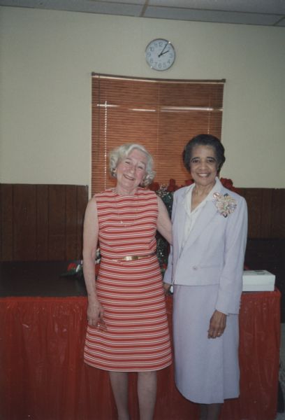 Vel Phillips posing with photographer Gerda Ruecktenwald. Vel is wearing a light blue suit, a flower shaped brooch, and a button pin that reads: "Hate" with a red slash through it. Gerda is wearing a striped red dress. Behind them on a table is a vase of red roses.