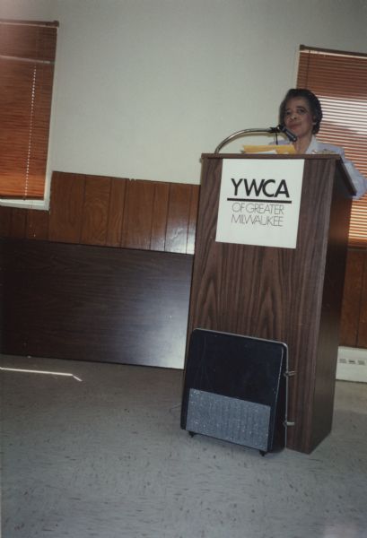 Vel Phillips speaking at a podium. She is wearing a light blue suit. The sign on the podium reads: "YWCA of Greater Milwaukee."