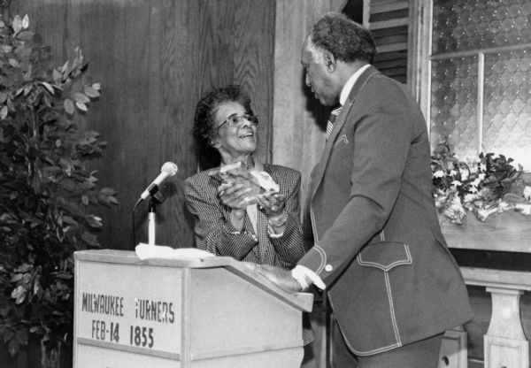 Vel Phillips, wearing a checkered suit and holding a glass award next to a podium. A man wearing a suit stands next to her. The sign on the podium reads: "Milwaukee Turners, Feb-14 1855."