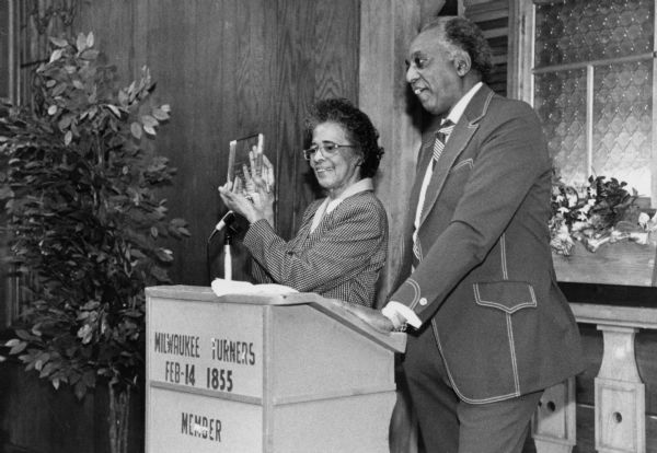 Vel Phillips, wearing a checkered suit is displaying a glass award while standing next to a podium. A man wearing a suit stands next to her. The sign on the podium reads: "Milwaukee Turners, Feb-14 1855, Member."