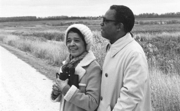 Vel Phillips, wearing a coat and a hat, stands on a gravel road with her husband, W. Dale Phillips, who is wearing a coat and eyeglasses. The marsh is in the background.