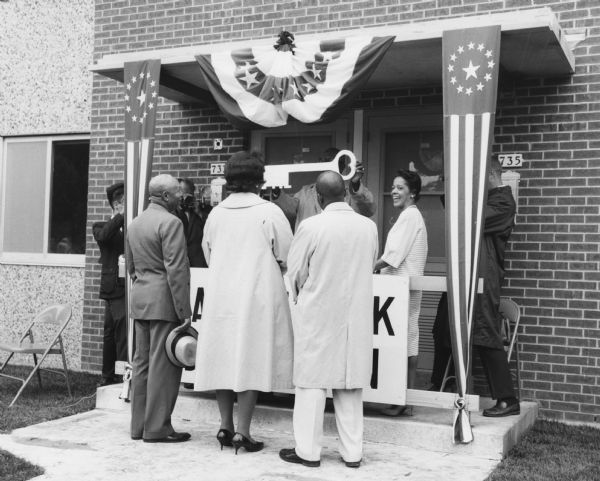 View of Mr. and Mrs. Paul Washington, standing on the sidewalk in the foreground with their backs to the camera, who were the first tenants of the Lapham Park Housing Project. They are being presented with a ceremonial key from a man standing behind a podium under the covered entrance to the building. Vel Phillips stands on the right wearing a striped dress. Photographers stand on the left and right side.