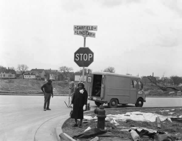 Bernice Lindsay is standing under a new reflected street name sign in her honor. She is wearing a coat, a hat, and a broach. Beside her is a fire hydrant. Behind her are two unidentified men. One is handling a tripod. There is a truck on the right and houses in the far background. Part of the N Lindsay Street Dedication ceremony.
