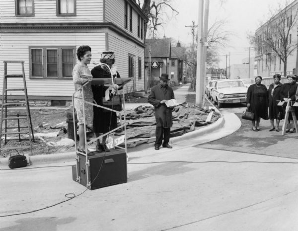 Vel Phillips is in a dress on a podium. She is next to Bernice Lindsay, who is wearing a coat and a hat. Behind them is Reverend Lovell Johnson, who is wearing a coat and a hat. He is holding papers in his hands. On the right are a group of unidentified women in coats. They are standing next to a street barricade. In the far background are trees, parked automobiles, and houses. Part of the N Lindsay Street Dedication ceremony.