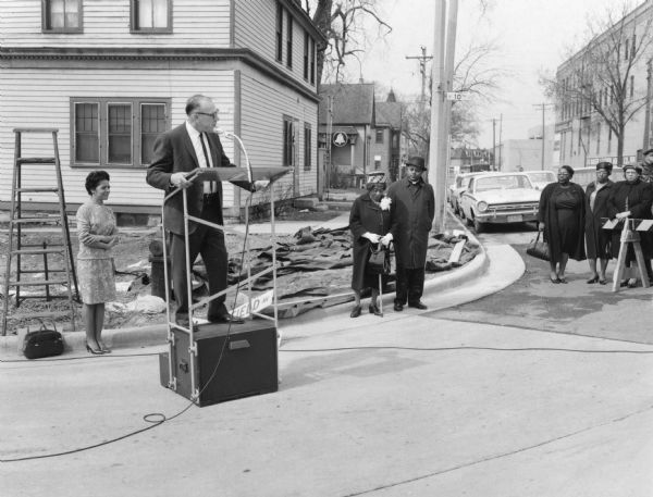 Alderman Martin E. Schreiber, President of the Milwaukee Common Council, is on a podium wearing a suit. On the left is Vel Phillips who is wearing a dress. On the right is Bernice Lindsay and Reverend Lovell Johnson. To the far right are a group of unidentified women standing next to a street barricade. In the background are automobiles, houses, and trees. Part of the N Lindsay Street Dedication ceremony.