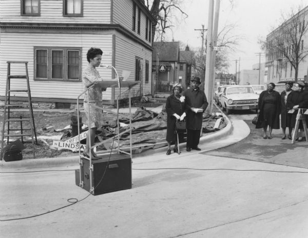6th Ward Milwaukee Alderman Vel Phillips is wearing a dress and standing on a podium. To the right are Bernice Lindsay and Reverend Lovell Johnson standing beside one another. To the far right are a group of unidentified women standing next to a street barricade. In the background are trees, automobiles, and houses. Part of the N Lindsay Street Dedication ceremony.