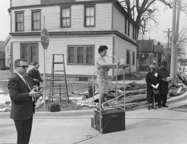 Vel Phillips is on a podium wearing a dress. To the left are two members of the media holding recording equipment who are documenting the N Lindsay Street Dedication ceremony. On the right are Bernice Lindsay and Reverend Lovell Johnson standing beside one another. In the background are trees, automobiles, and houses.