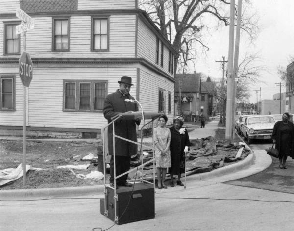 Reverend Lovell Johnson of St. Marks African Methodist Episcopal Church is on a podium in a coat and hat. On her right are Vel Phillips and Bernice Lindsay standing next to each other. On the far right is an unidentified woman. In the background are automobiles, trees, and houses. Part of the N. Lindsay Street Dedication ceremony.
