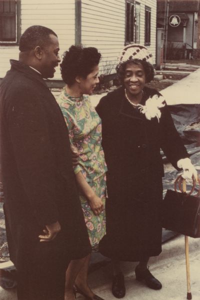 Reverend Lovell Johnson is on the left wearing a coat. On the right is Bernice Lindsay who is wearing a hat and carrying a handbag and cane. In the middle is Alderman Vel Phillips, who is wearing a colorful dress. Part of the N Lindsay Street Dedication ceremony.