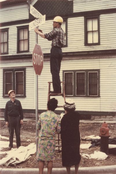 Vel Phillips is wearing a colorful dress. On the right is Bernice Lindsay, who is wearing a coat and a hat. Both are looking at a worker who is installing the N Lindsay Street sign on a ladder. There is another worker on the left looking up. There is a fire hydrant in the bottom right corner. Part of the N Lindsay Street Dedication ceremony.