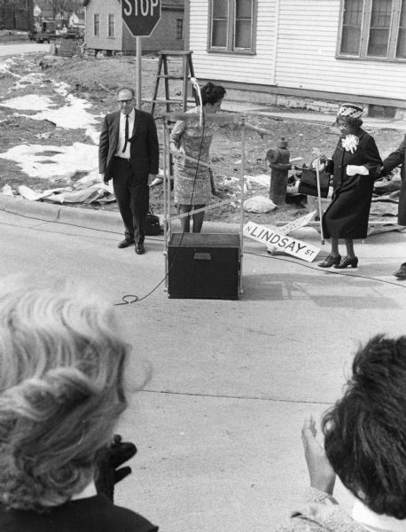 Vel Phillips is behind the podium in a dress. She is reaching out her hand for Bernice Lindsay, who is wearing a coat and a hat. An unidentified person is helping her walk. On the left is Alderman Martin Schreiber, who is wearing a suit and glasses. Two unidentified women are in the foreground applauding.