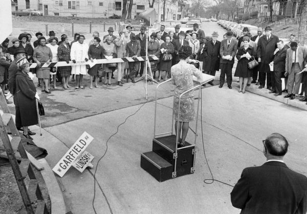 Vel Phillips is at a podium speaking to a crowd of people, some of whom are standing behind a street barricade. She is wearing a dress. In the bottom right corner is Alderman Martin Schreiber. He is wearing a suit and glasses. On the left is Bernice Lindsay, who is wearing a coat and a hat. She is carrying a cane. Next to her is Reverend Lovell Johnson. He is wearing a hat and a coat. In the background are trees, automobiles, and houses. Part of the N Lindsay Street Dedication ceremony.