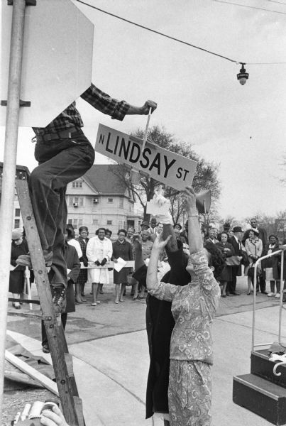 Vel Phillips and Bernice Lindsay helping lift the Lindsay Street sign to be installed. Phillips is wearing a dress and is in front of Lindsay, who is wearing a dark coat and a hat. There is an unidentified worker on a ladder holding onto the sign. In the bottom left corner is someone shooting footage of the event. Reverend Lovell Johnson is behind the worker and is wearing a hat and coat. There is a crowd in the background. Behind the crowd are trees and houses.