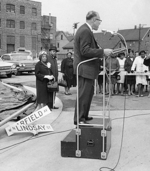 President of the Milwaukee Common Council Martin E. Schreiber is at the podium. He is wearing a suit and glasses. On the left is Bernice Lindsay who is wearing a hat and holding a cane and handbag. Next to her is Reverend Lovell Johnson. He is wearing a coat and a hat. In the background is a crowd of people behind a street barricade. Behind the crowd are houses and automobiles.