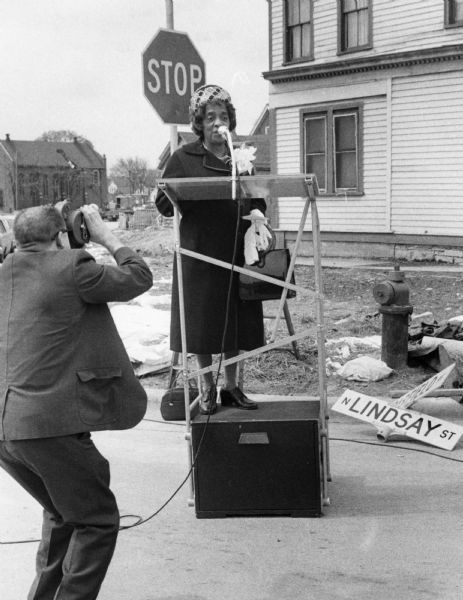 Bernice Lindsay is on a podium. She is wearing a coat and a hat. A man is filming her speech. On the ground is the street sign in her honor. Part of the N Lindsay Street Dedication ceremony.