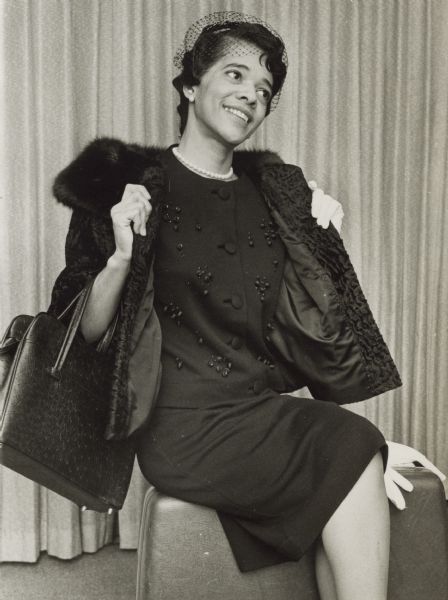 Portrait of Vel Phillips sitting and posing for a fashion shoot. She is wearing a dark outfit with a fur-lined coat. She has a handbag on her right arm and is wearing a pearl necklace.