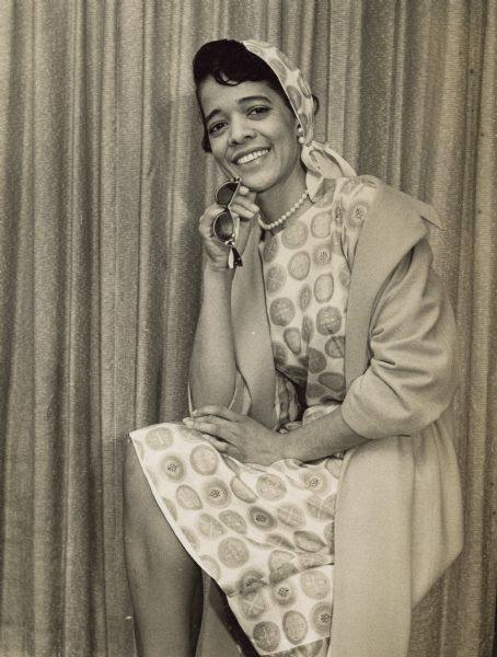 Portrait of Vel Phillips sitting and posing. She is wearing a coat over a dress and matching headscarf, and is holding sunglasses in her hand.