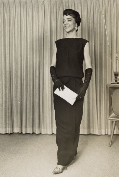 Full-length portrait of Vel Phillips wearing a dark dress with matching elbow-length gloves and earrings. She is holding a light-colored clutch handbag. In the background is a television.