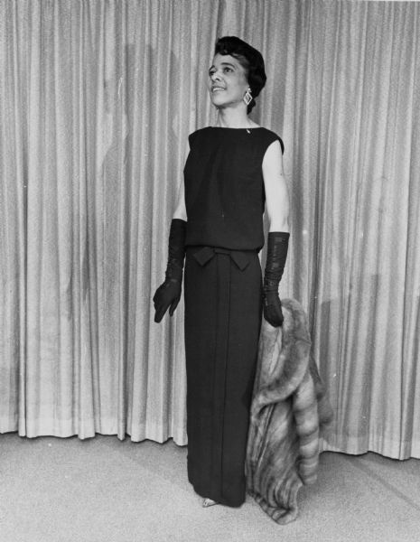Full-length portrait of Vel Phillips wearing a dark dress. She is also wearing earrings and elbow length gloves, and is holding a fur coat in her left hand.