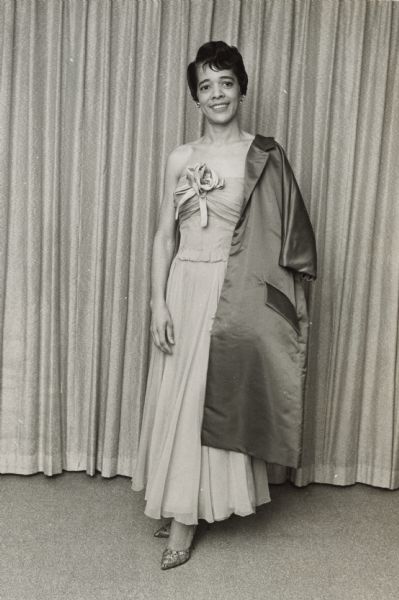 Full-length portrait of Vel Phillips wearing a strapless dress with a flower detail in the center. She is wearing heels and has a satin coat draped over her left shoulder.