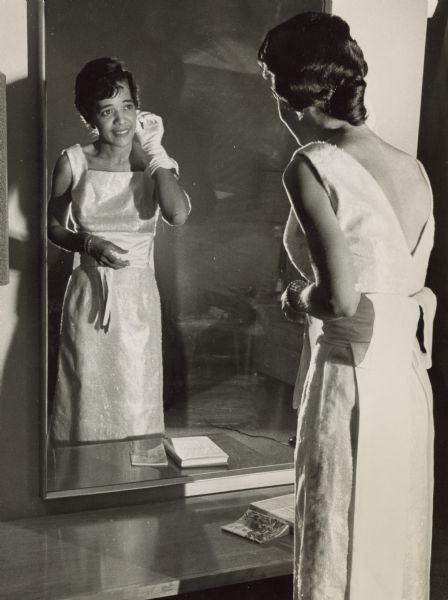 Vel Phillips wearing a light-colored dress. She is wearing a white glove on her right hand and has bracelets on her left wrist. She is looking in a mirror. There are books on the counter in front of her.